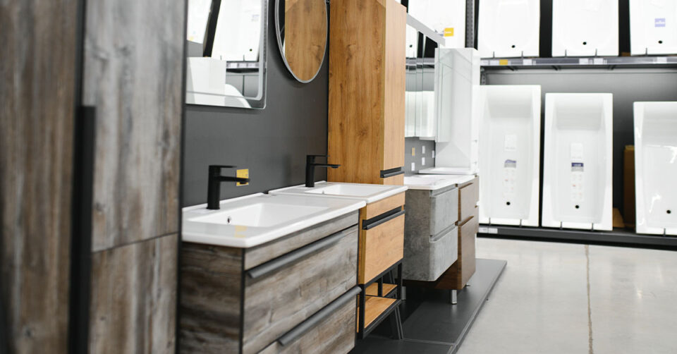 Display of contemporary bathroom vanities with wood and stone finishes, black faucets, and white sinks in a retail showroom.