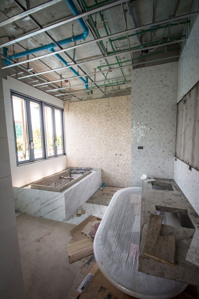 Bathroom under construction with exposed ceiling pipes, unfinished tile work, and fixtures still being installed.