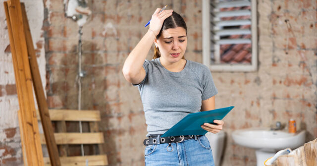 Frustrated woman holding a clipboard and pen in an unfinished bathroom under renovation.