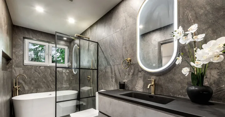 Contemporary bathroom with dark stone walls, a freestanding tub, glass shower enclosure, gold fixtures, and an illuminated oval mirror.