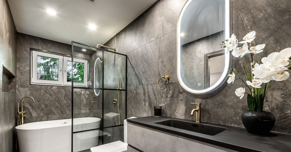 Contemporary bathroom with dark stone walls, a freestanding tub, glass shower enclosure, gold fixtures, and an illuminated oval mirror.