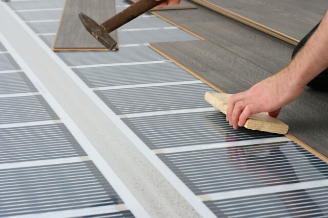 Person installing laminate flooring over an electric radiant floor heating system using a tapping block and hammer.