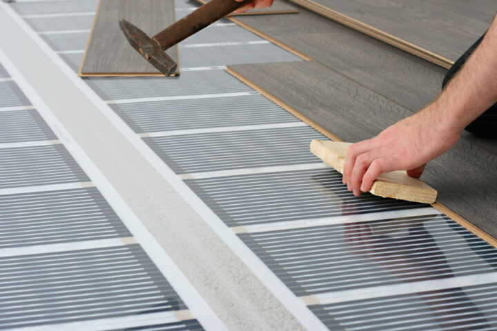 Person installing laminate flooring over an electric radiant floor heating system using a tapping block and hammer.