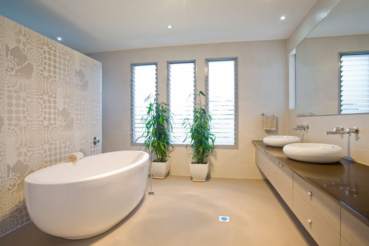 Modern beige bathroom featuring a freestanding bathtub, dual vessel sinks on a dark countertop, potted plants, and decorative wall tiles.