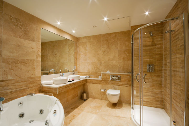 Luxurious bathroom featuring beige marble tiles, a whirlpool bathtub, wall-mounted toilet, dual sink vanity, and a curved glass shower enclosure.