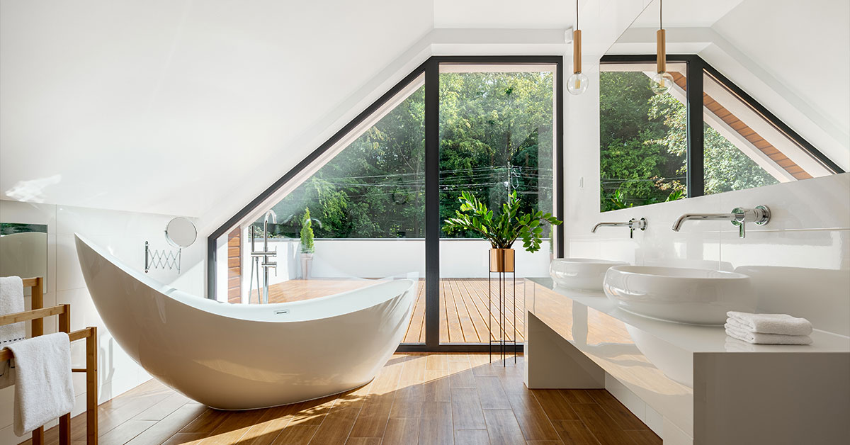 Bright attic bathroom with a sculptural freestanding tub, double vessel sinks, and large triangular windows overlooking greenery.