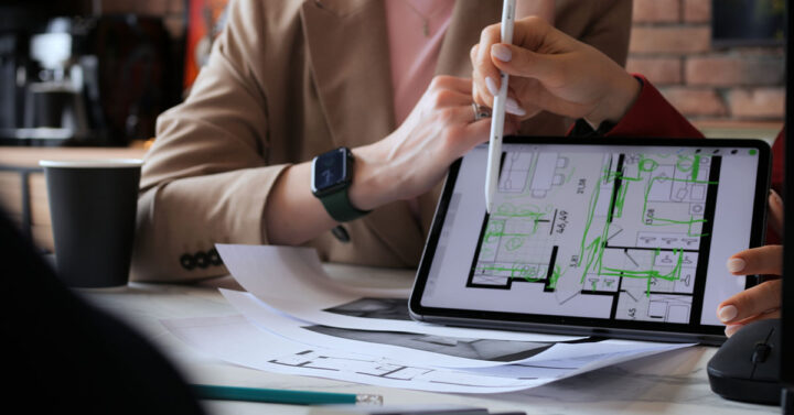 Close-up of two people reviewing and editing architectural floor plans on a tablet with printed blueprints on the table.
