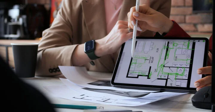 Close-up of two people reviewing and editing architectural floor plans on a tablet with printed blueprints on the table.