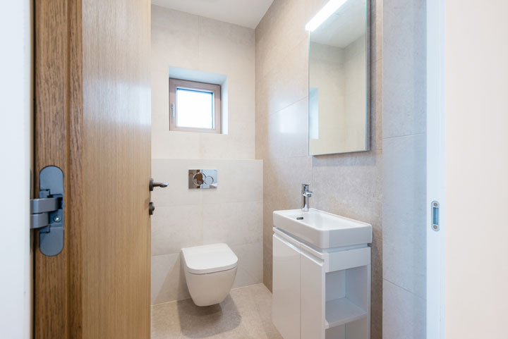 Compact modern bathroom with a wall-mounted toilet, small vanity with sink, and a square window for natural light.