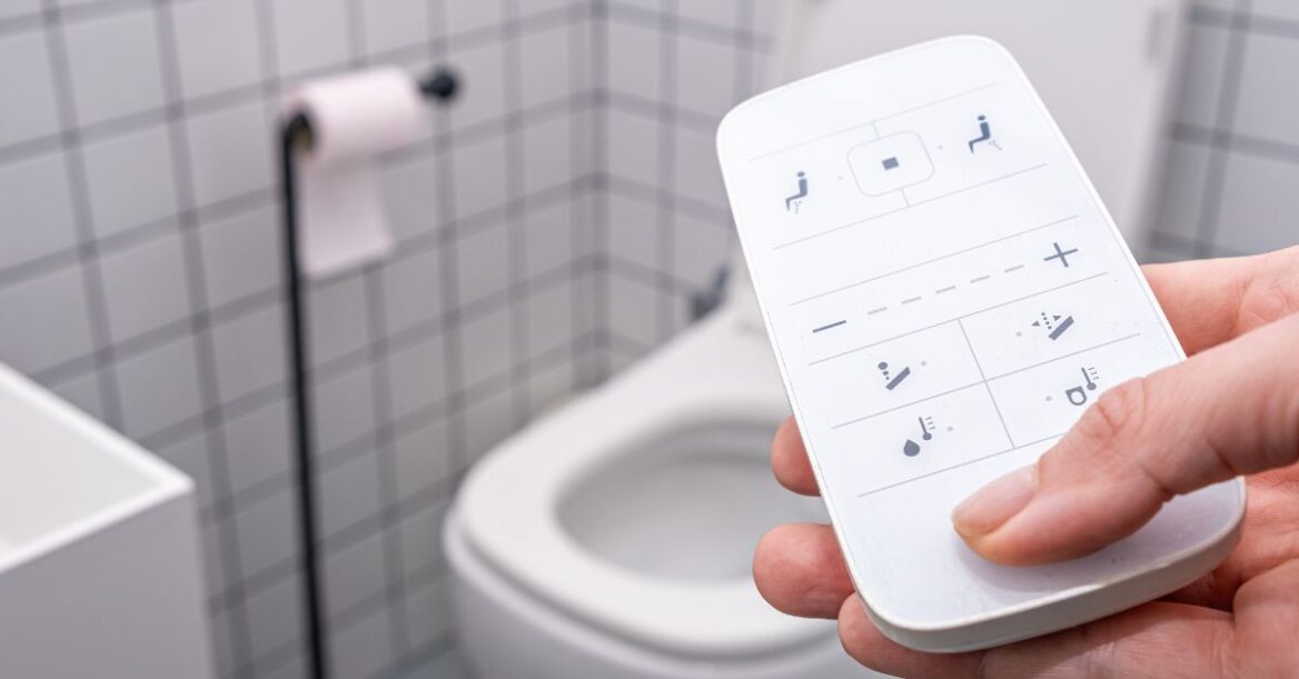 Person holding remote control that controls a smart toilet which is in the backgroud.