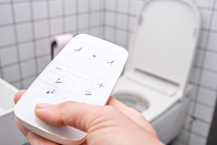 Close-up of a person holding a remote control for a smart toilet in a tiled bathroom.