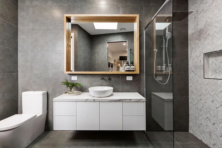 Modern bathroom with dark gray tiles, a white floating vanity, vessel sink, walk-in glass shower, and a large framed mirror.